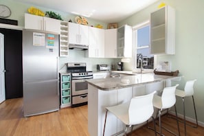 A modern kitchen with white cabinets, a stainless steel refrigerator, an oven, a stove, a coffee maker, and a window with a view of a house. - Oceanfront 1Br/1Ba condo~ The One (Folly Beach)