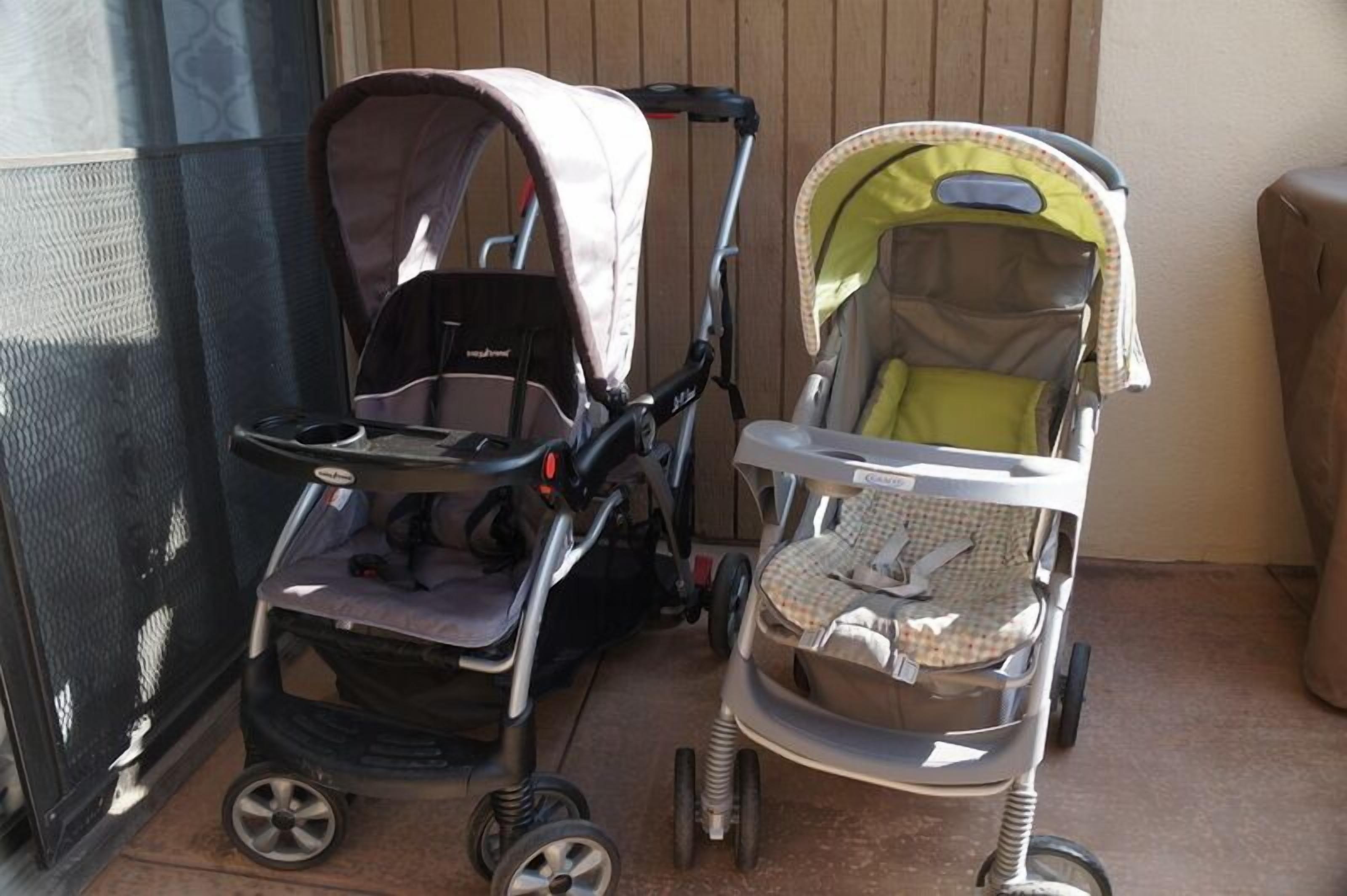 Two strollers parked on a tiled patio next to a wooden fence.