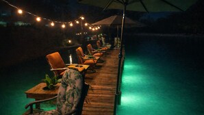 Zona junto a la piscina con sillones y mesas de madera, luces colgantes y sombrillas.
