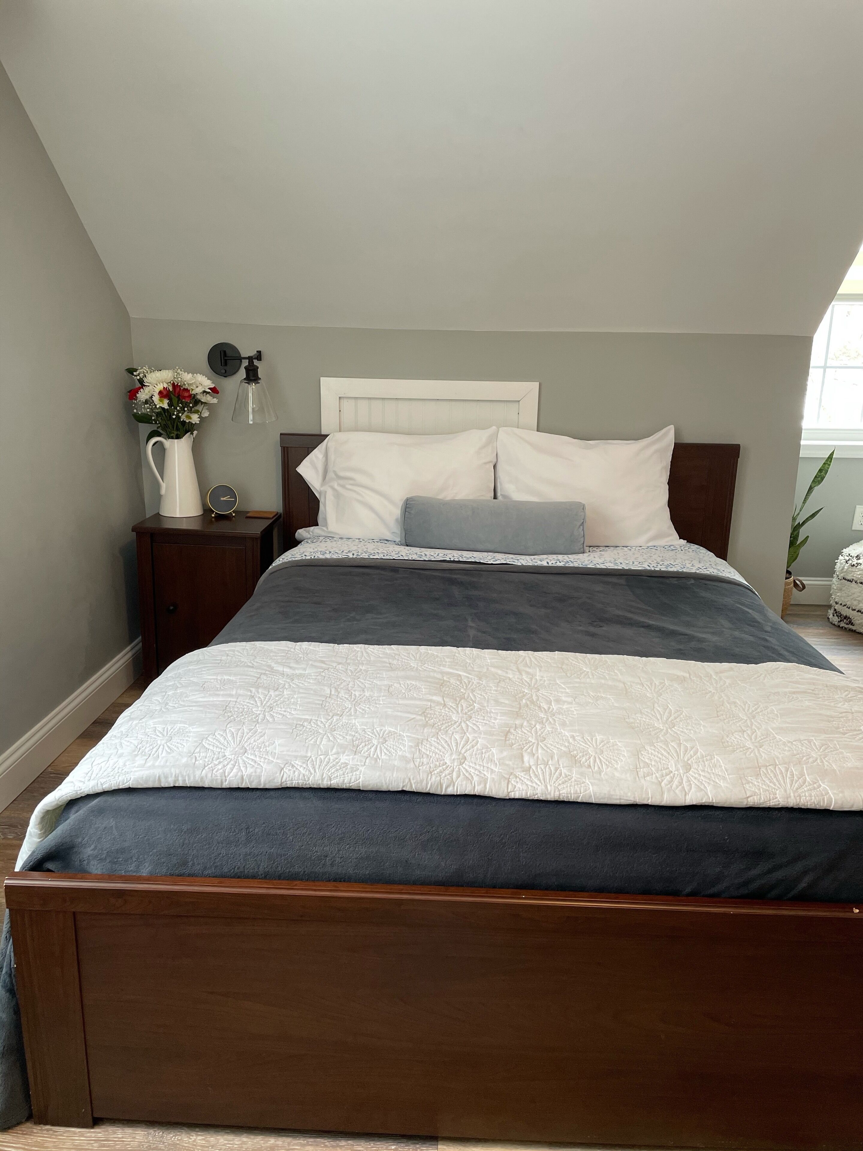 A bedroom with a wooden bed, white and gray bedding, a nightstand with a vase of flowers, a wall clock, and a window with a view of a plant.
