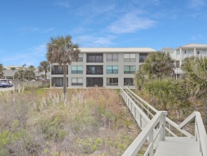 A modern multi-story building with a flat roof, surrounded by palm trees and dry grass. - Oceanfront condo with beautiful ocean view and screened patio- SO 4 (St. Augustine Beach)