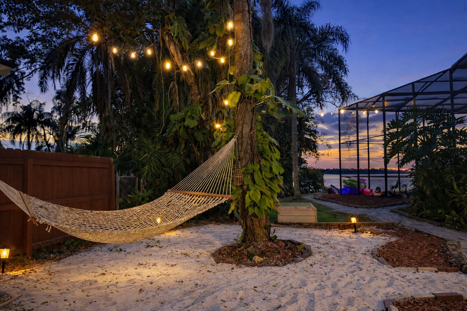 A hammock tied to a tree in a sandy area with string lights, surrounded by greenery and a glass structure.