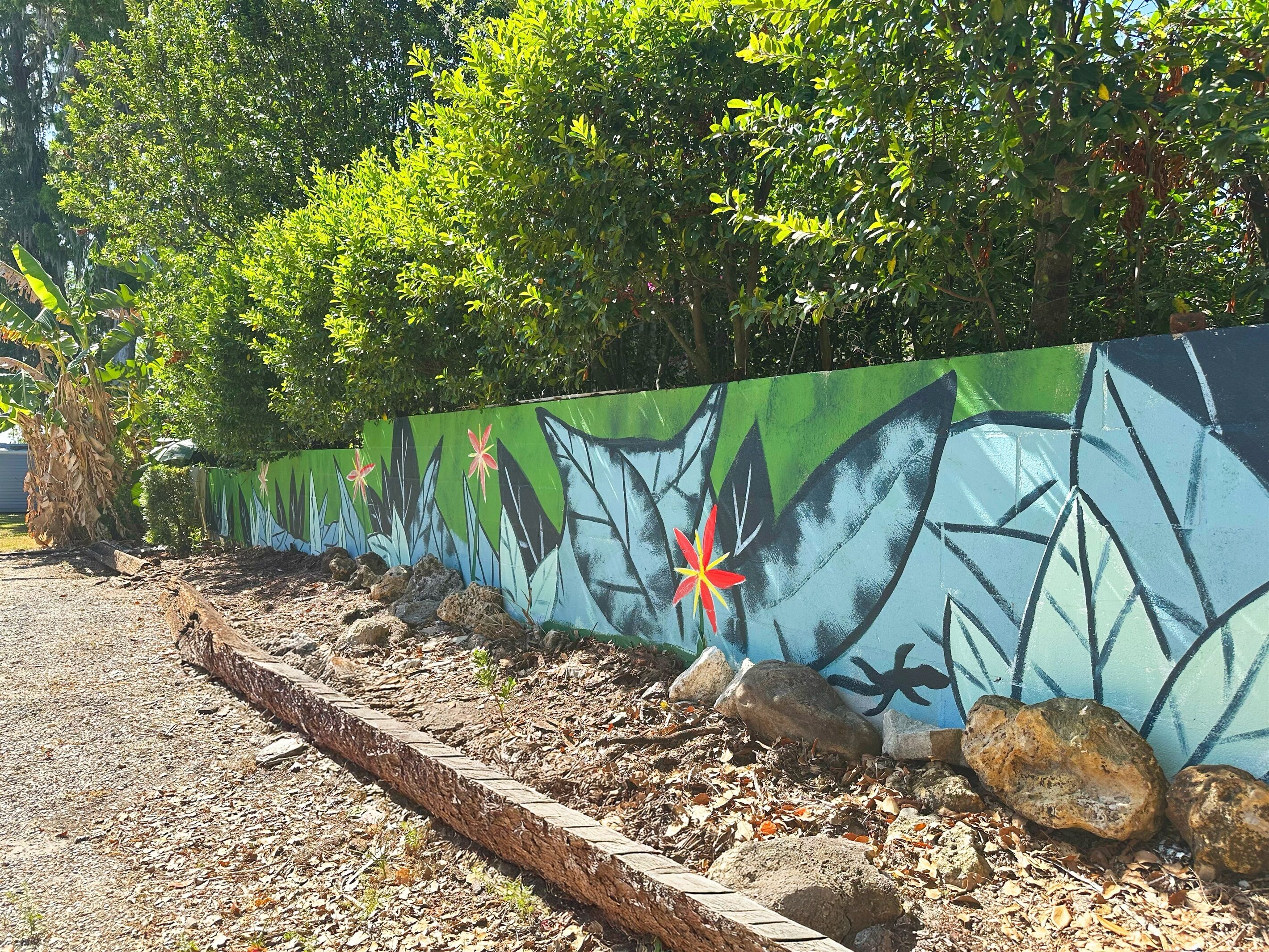A garden area with a painted wall featuring tropical plants and flowers, surrounded by rocks and a wooden border.