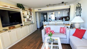 Un salon avec un canapé gris, des coussins rouges, une table basse avec des livres et un vase, un téléviseur sur le côté gauche et une cuisine à l’arrière-plan.