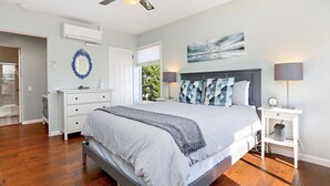 Une chambre à coucher comprenant un lit, des tables de chevet, un ventilateur de plafond, un climatiseur mural, une commode et une fenêtre donnant sur un paysage verdoyant.