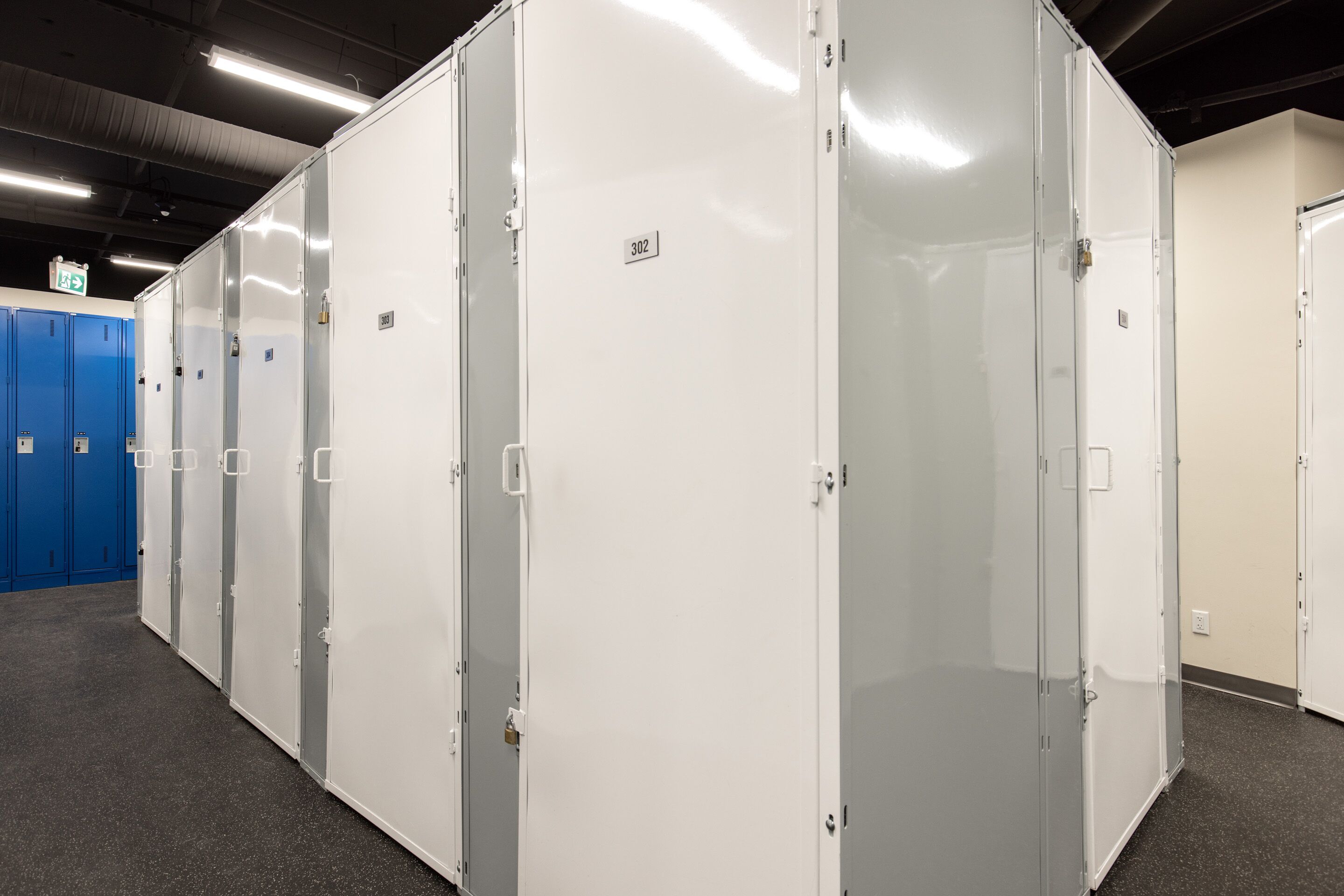A row of white lockers with a number tag, blue lockers in the background, and a carpeted floor.