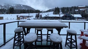 Zona salotto esterna coperta di neve con tavolo e sgabelli, affacciata su una località sciistica con edifici e una seggiovia.