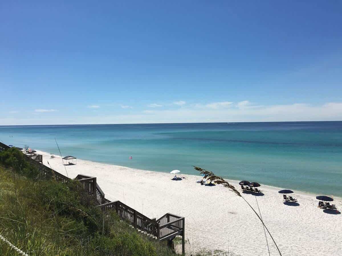 Une plage avec une promenade en bois menant au bord de l’eau, une eau d’un bleu limpide et des parasols de plage.