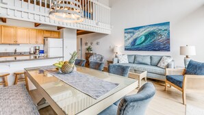 A spacious living area with a glass-top dining table, a high ceiling with wooden beams, a white railing, and a large abstract painting on the wall. - Rare, Spacious Penthouse, Big Ocean and Mountain Views, Great Location,  AC (Maui)