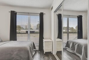 Two beds with grey bedding, a nightstand, and a large mirror in a stylish room with a window offering a view of the surrounding area.