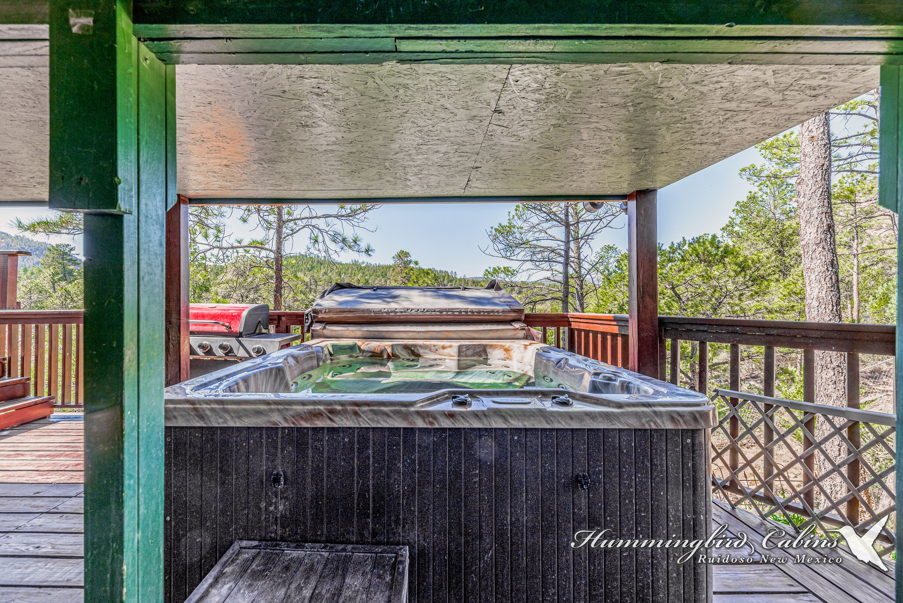A hot tub under a wooden deck with a view of trees and a grill in the background.