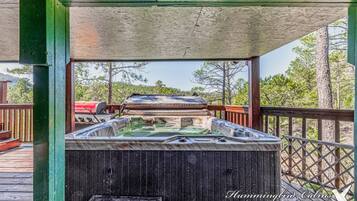 A hot tub under a wooden deck with a view of trees and a grill in the background.