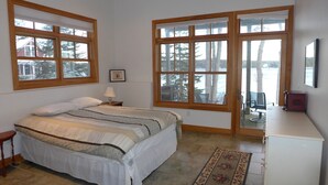 Ein Schlafzimmer mit einem Bett, einem Nachttisch, einer Kommode und großen Fenstern mit Blick auf Bäume und ein Gebäude.