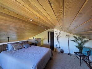 Un dormitorio con techo inclinado, una cama, lámparas de noche, una silla, una planta y un aplique de pared.