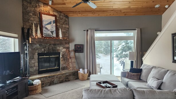 A cozy living room with a stone fireplace, a ceiling fan, and a large window with a view of snow-covered trees. - Cozy Big Sky Chalet - Perfect For Your Montana Getaway (Big Sky)