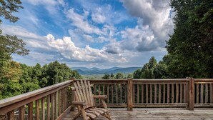 Una terraza de madera con una silla y una mesa de madera, con vistas a un área boscosa y montañas a lo lejos.