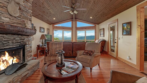 Un salón amplio con chimenea de piedra, techo de madera y ventanas grandes con vistas a la montaña.