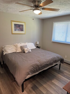 A bedroom with a bed, a ceiling fan, a framed picture on the wall, and a window with blinds.