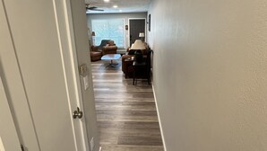 Ein Flur mit Holzboden, einer Tür links und einem Wohnbereich mit Couch und Lampe im Hintergrund.