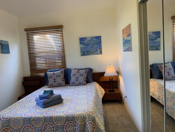 Une chambre à coucher avec un lit, une table de chevet avec une lampe, une fenêtre avec des stores et deux tableaux accrochés au mur.