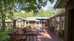 A wooden deck with picnic tables and chairs, a covered patio area, and a view of a cabin-style building. - 3-Bed Sleeping Cabin Near Geneva Lake – Walk to Dining & Beach (Williams Bay)
