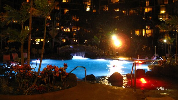 Vista nocturna de un área de piscina de hotel con cascada, palmeras y un puente.