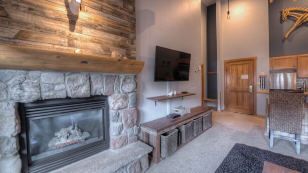 A stone fireplace with a television above it, a wooden bench, and a wicker chair. - Walk to Lifts! Free Covered Parking, High Ceilings (Dillon)