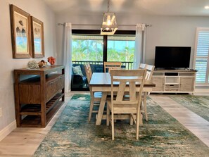 Un coin repas avec une table et des chaises en bois, un téléviseur posé sur un meuble, et une vue sur l’extérieur grâce à une porte coulissante.