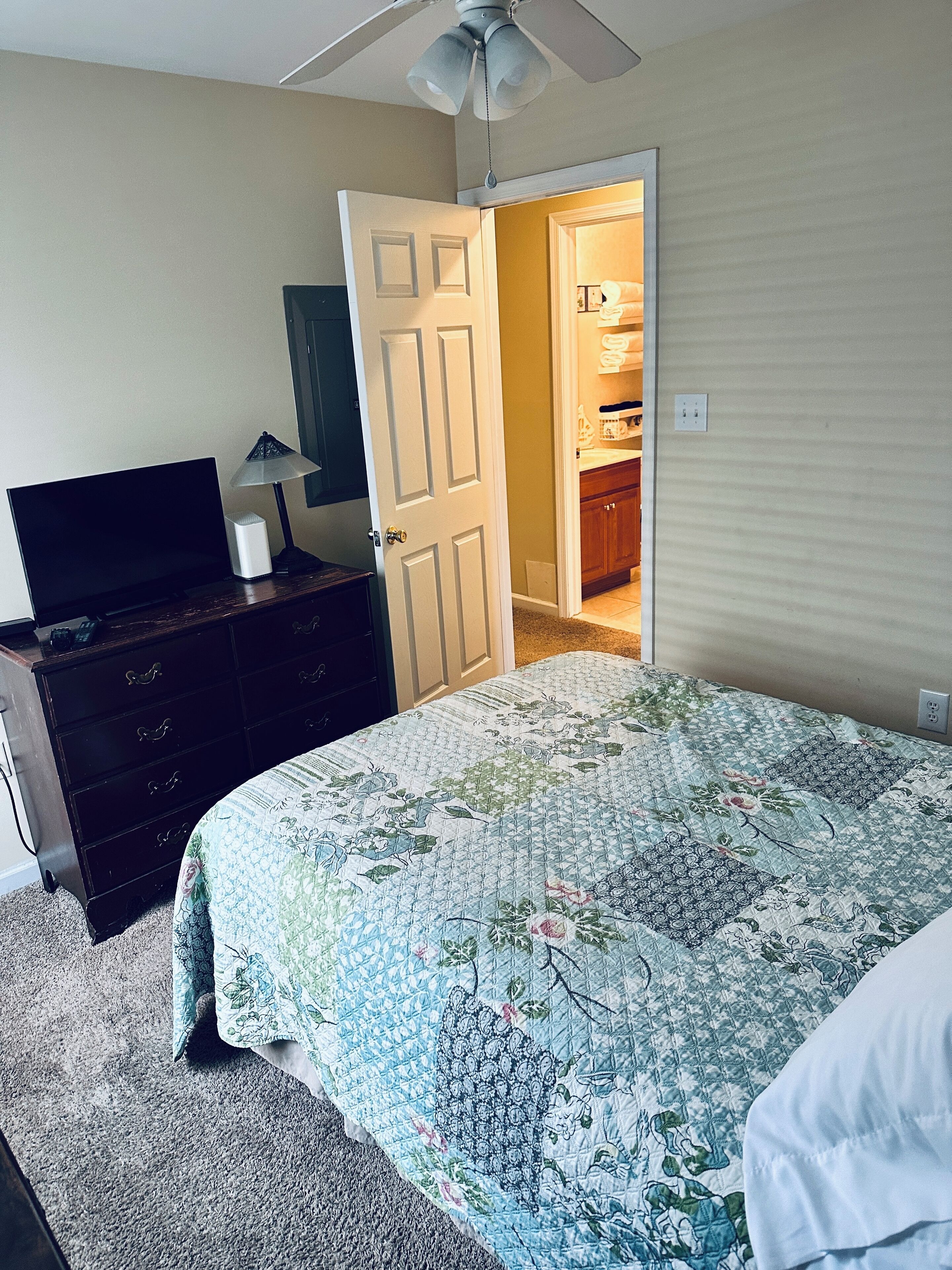 Une chambre à coucher comprenant un lit, une commode, un téléviseur et une porte ouverte donnant sur une salle de bain.