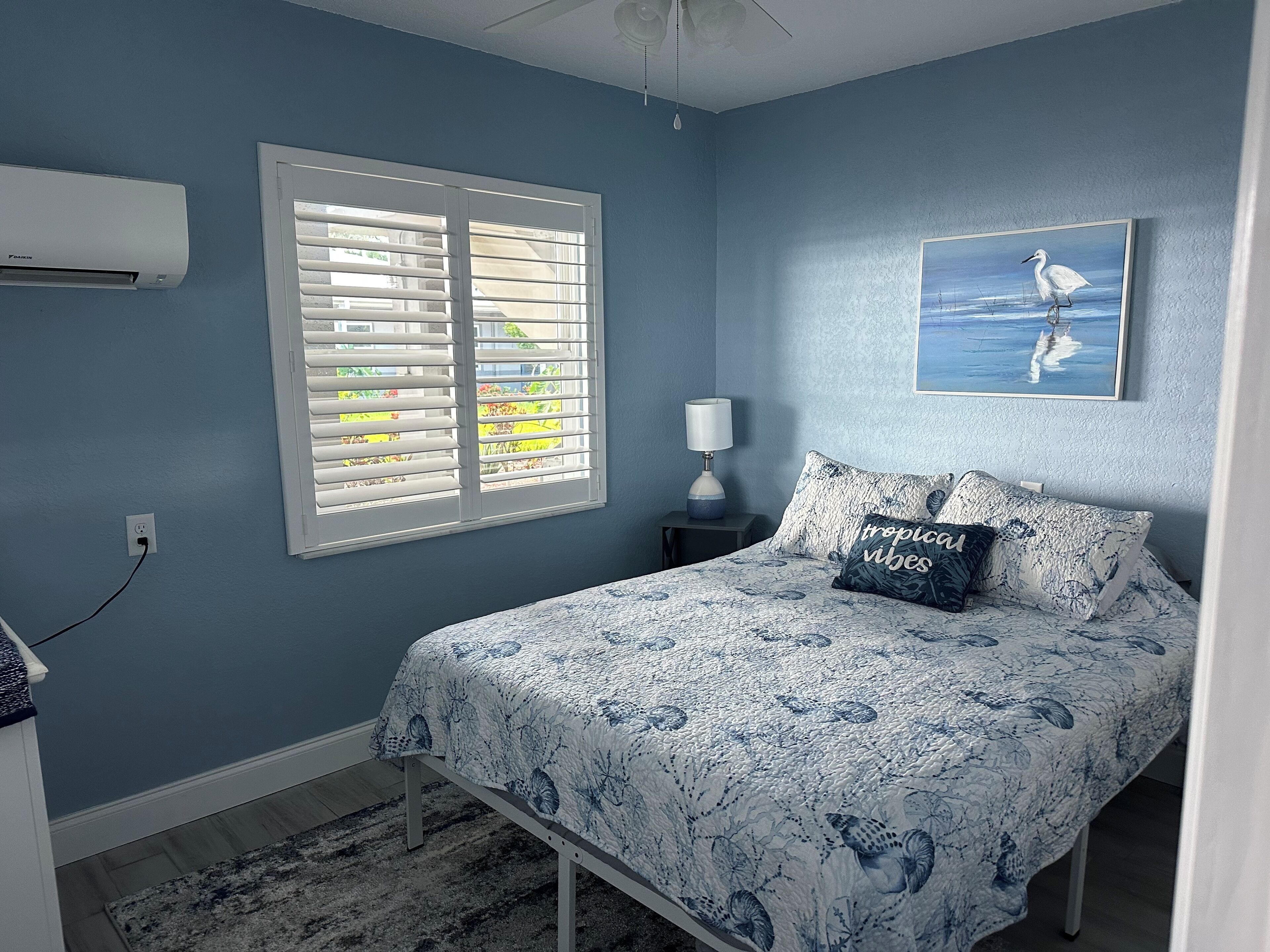 Un dormitorio con cama, ventana con persianas, aire acondicionado, mesita de noche con lámpara y un cuadro en la pared.