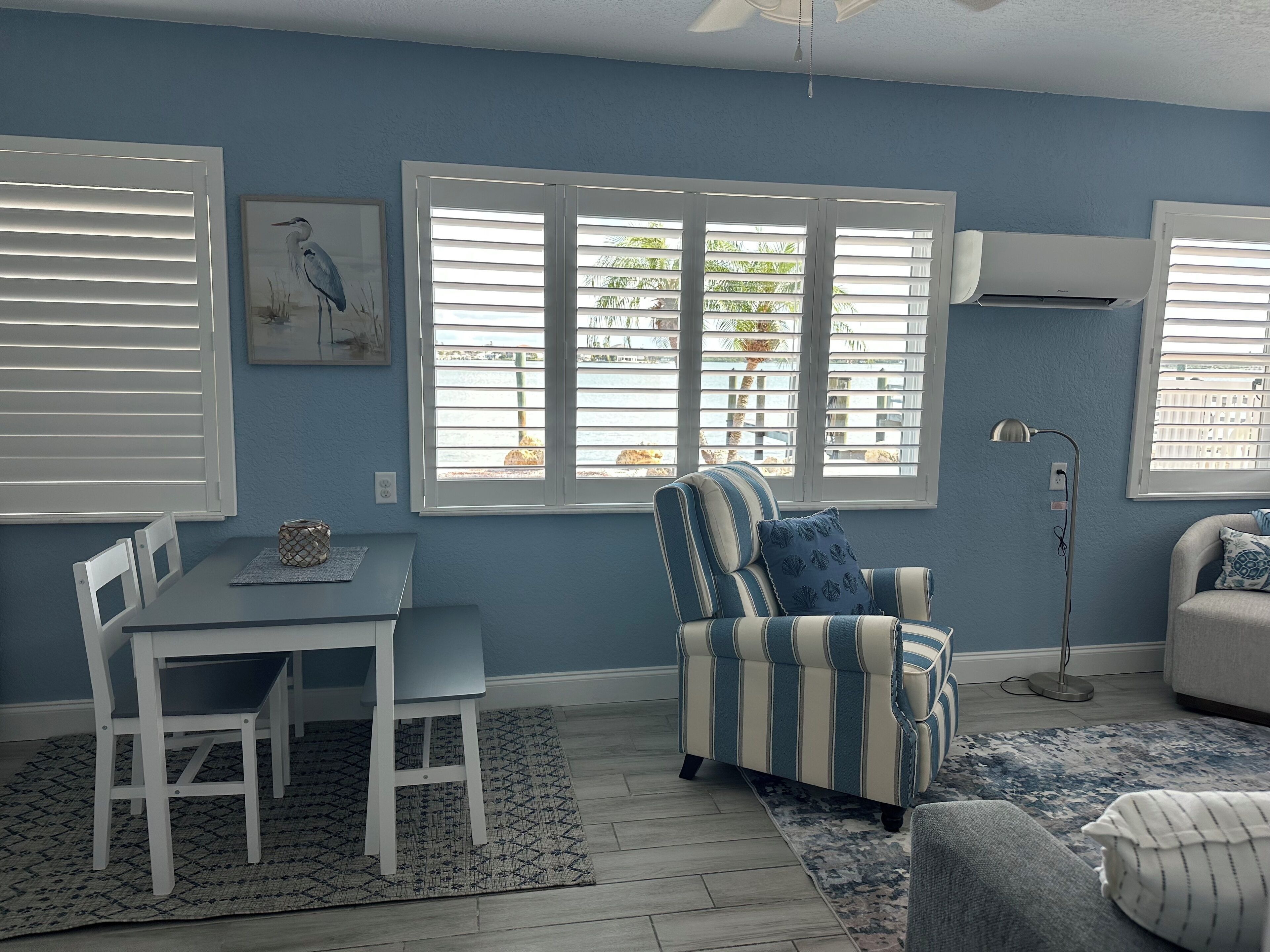 Ein Wohnzimmer mit einem gestreiften Sessel, einem Esstisch mit Stühlen, einer Stehlampe und einem Fenster mit Jalousien.