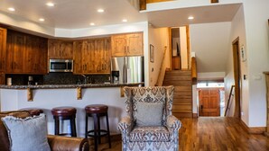 Une cuisine avec des meubles en bois, un four à micro-ondes et un réfrigérateur. On y trouve un fauteuil à motifs et trois tabourets de bar. Un escalier en bois mène à une porte située au fond de la pièce.