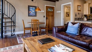A living room with a brown leather sofa, a wooden coffee table with magazines, a dining area with a wooden table and chairs, and a kitchen in the background.