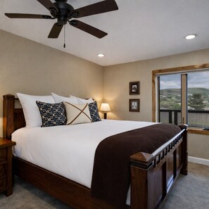 A bedroom with a large bed, a ceiling fan, a nightstand, a lamp, and a window with a view. - Quitting Time Ultimate Old Town Adventure Basecamp 447 (Park City)
