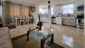 Una cocina moderna con armarios blancos, horno empotrado y una zona de comedor con mesa y sillas de vidrio.