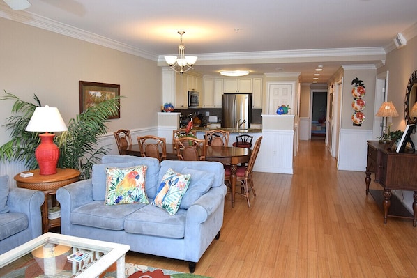 A spacious living room with a blue sofa, a coffee table, and a dining area with a wooden table and chairs. A kitchen with white cabinets and stainless steel appliances is visible in the background.