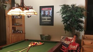 Una mesa de billar con tacos, una lámpara, un cartel de Budweiser y una planta en maceta.