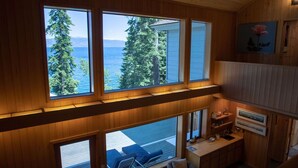 A cabin with large windows overlooking a forest and water, a cozy seating area, and wooden interior design.