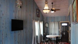 Una habitación con paredes de madera, ventilador de techo, candelabro, televisor de pantalla plana, mesa de comedor con sillas y sofá rojo.