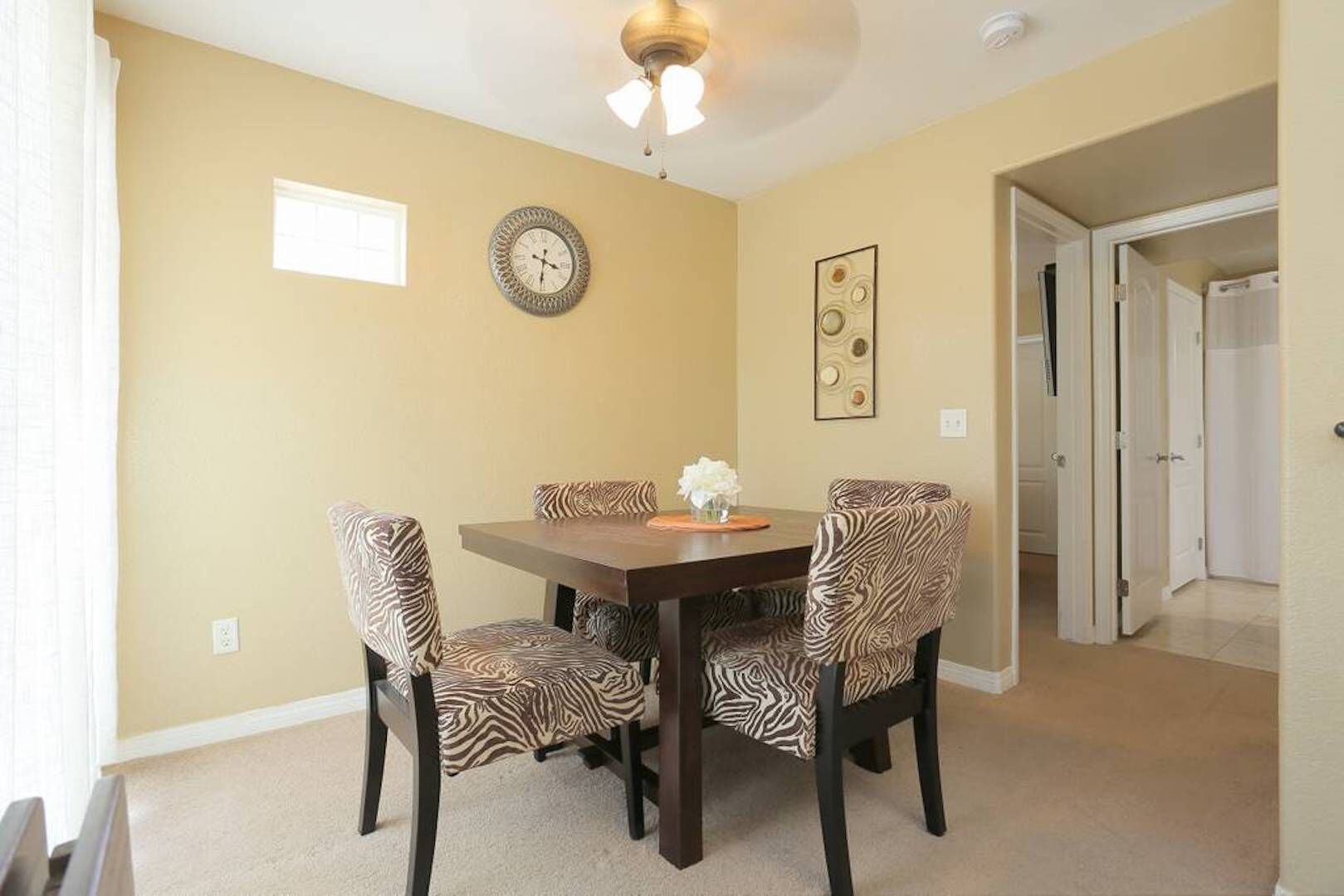 A dining room with a wooden table and zebra-patterned chairs, a wall clock, and a ceiling fan.
