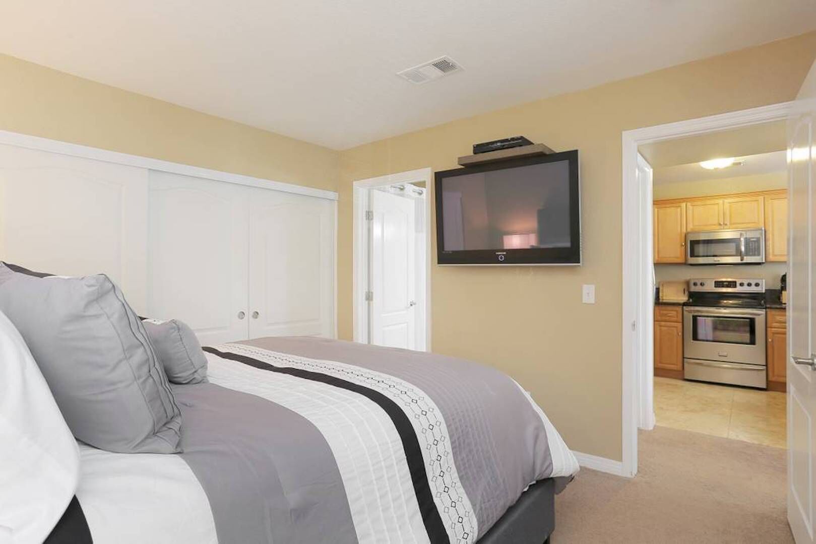 Un dormitorio con una cama, un televisor montado en la pared y una cocina visible a través de una puerta abierta.