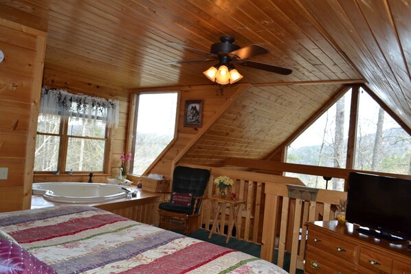 A cozy cabin with a large bed, a ceiling fan, a television, and a bathtub.