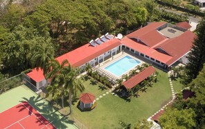 Vue aérienne d’un complexe hôtelier doté d’une piscine, de courts de tennis et d’une végétation luxuriante.