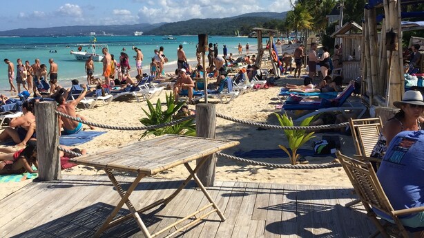Une terrasse en bois, dotée de chaises longues et de parasols, qui offre une vue sur une plage animée, avec des personnes et des palmiers.