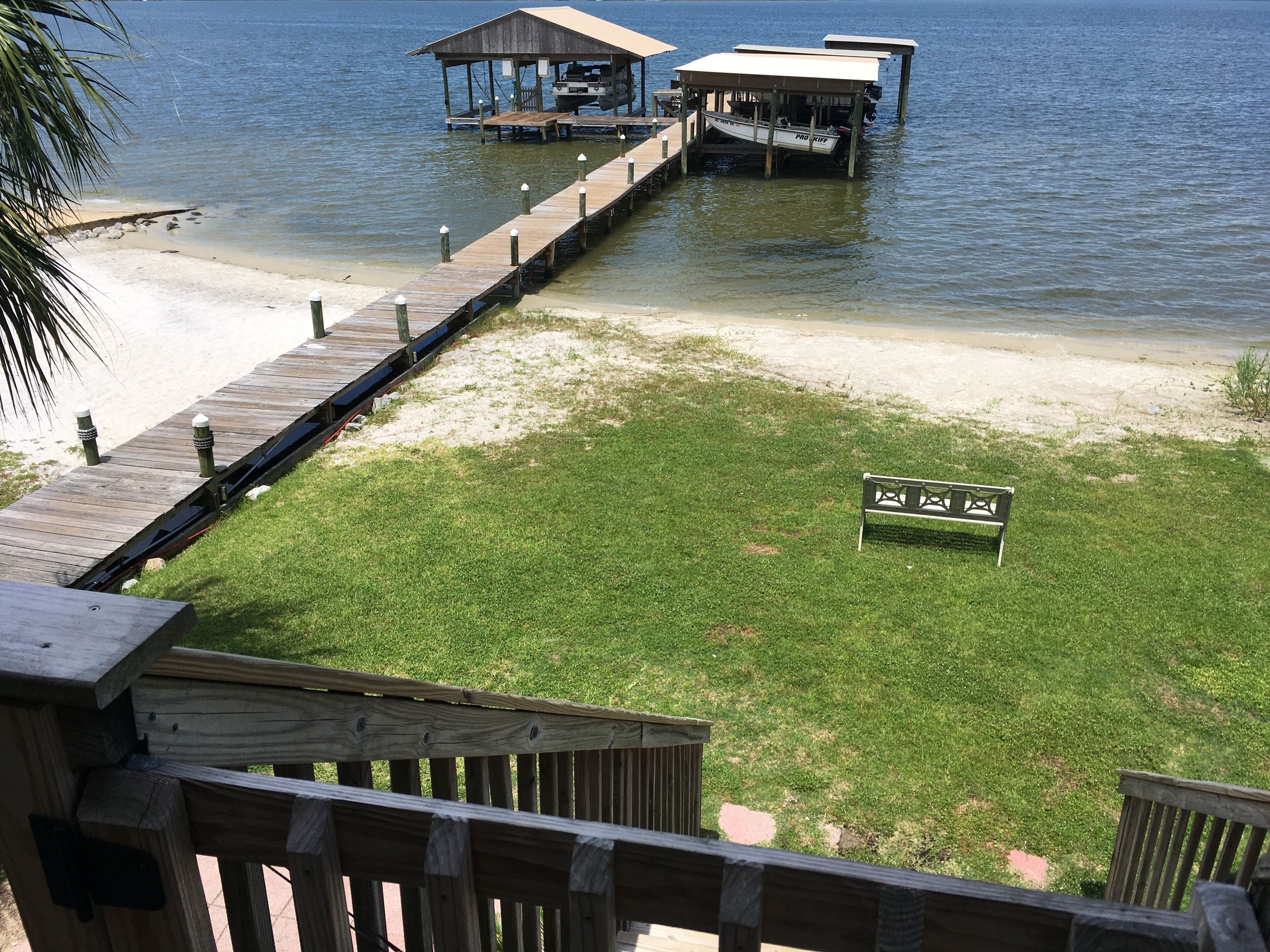 Blick auf einen Strand mit einer hölzernen Pier, die zu einem Anlegesteg führt, einer Rasenfläche und einem Schild, das Bootsverleih Pro Kiff anzeigt.