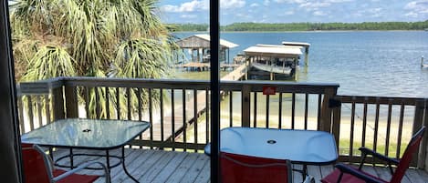 A wooden deck with a glass table and red chairs, overlooking a body of water with boats and a dock.