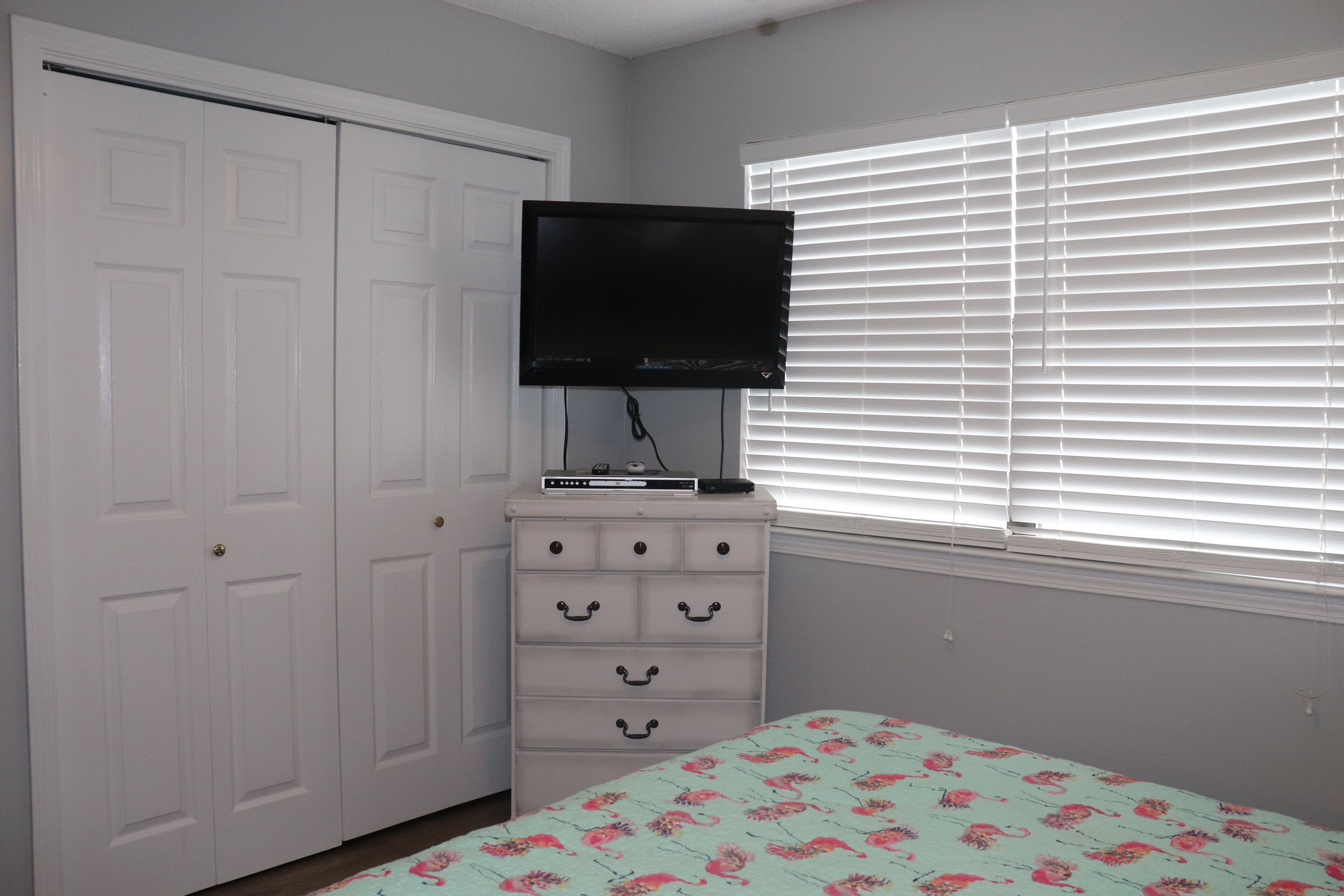 A bedroom with a bed, a dresser, a television, and a window with blinds.
