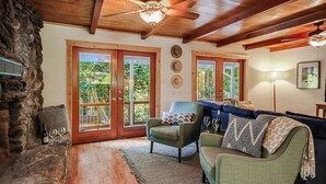 A cozy living room with a stone fireplace, wooden ceiling beams, and a large window offering a view of greenery. - Relaxing Retreat in the Redwoods, Spa, Dog Friendly! (Cazadero)