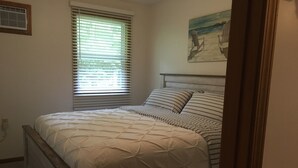 Un dormitorio con cama, ventana con persianas, un cuadro en la pared y cabecero de madera.