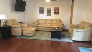 Una sala de estar con un sofá color crema, dos sillones, una mesa de centro, un tapete estampado, un televisor de pantalla plana y un ventilador de techo.